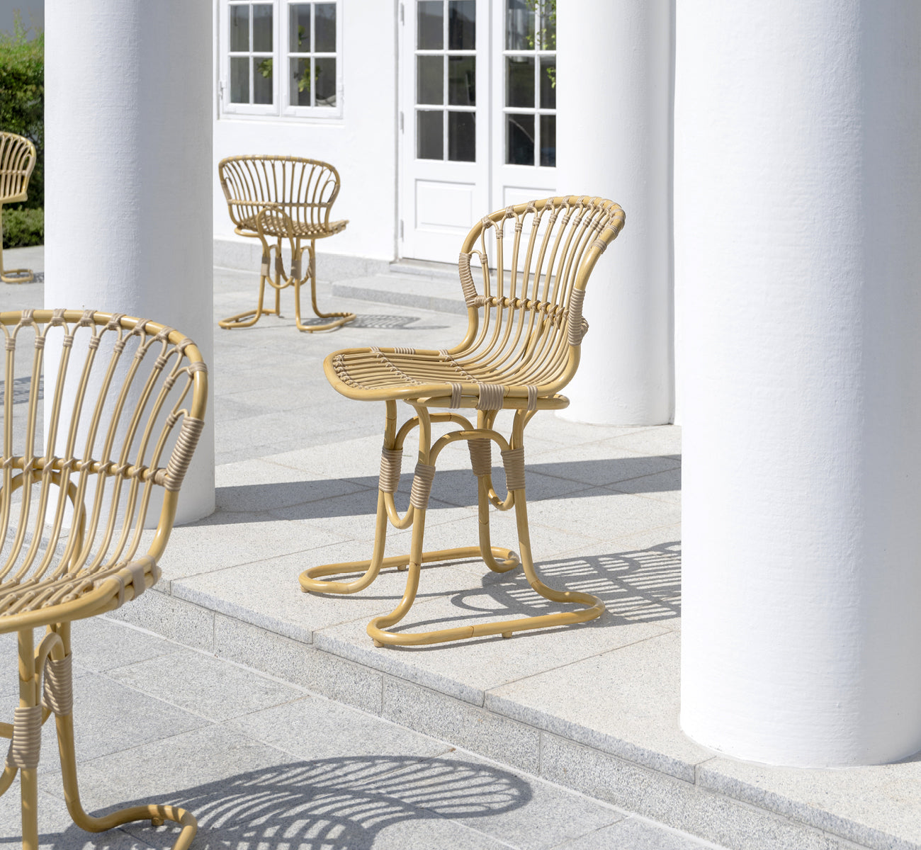 This is a lifestyle image of Rita Exterior Dining Chair in natural aluminium artfibre. Paired with a bistro table on a white colonnaded terrace.