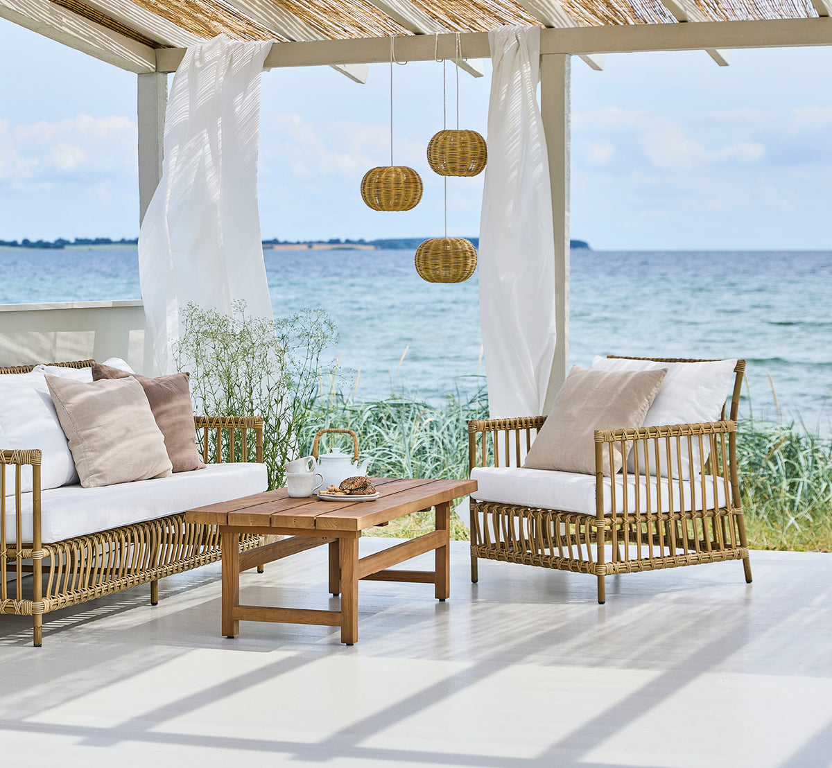 This is a lifestyle image of Caroline Exterior Lounge Chair in antique Aluminium ArtFibre, on a waterside wooden deck with coastal greenery and sea.