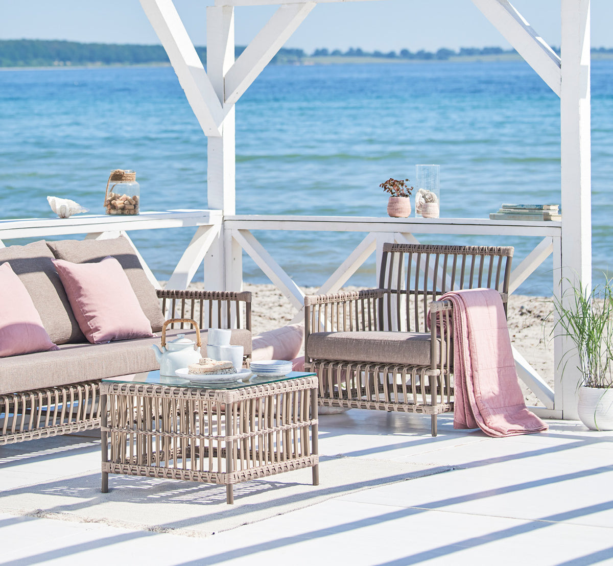 This is a lifestyle image of Caroline Exterior Lounge Chair in moccaccino Aluminium ArtFibre, on a waterside wooden deck with coastal greenery and.