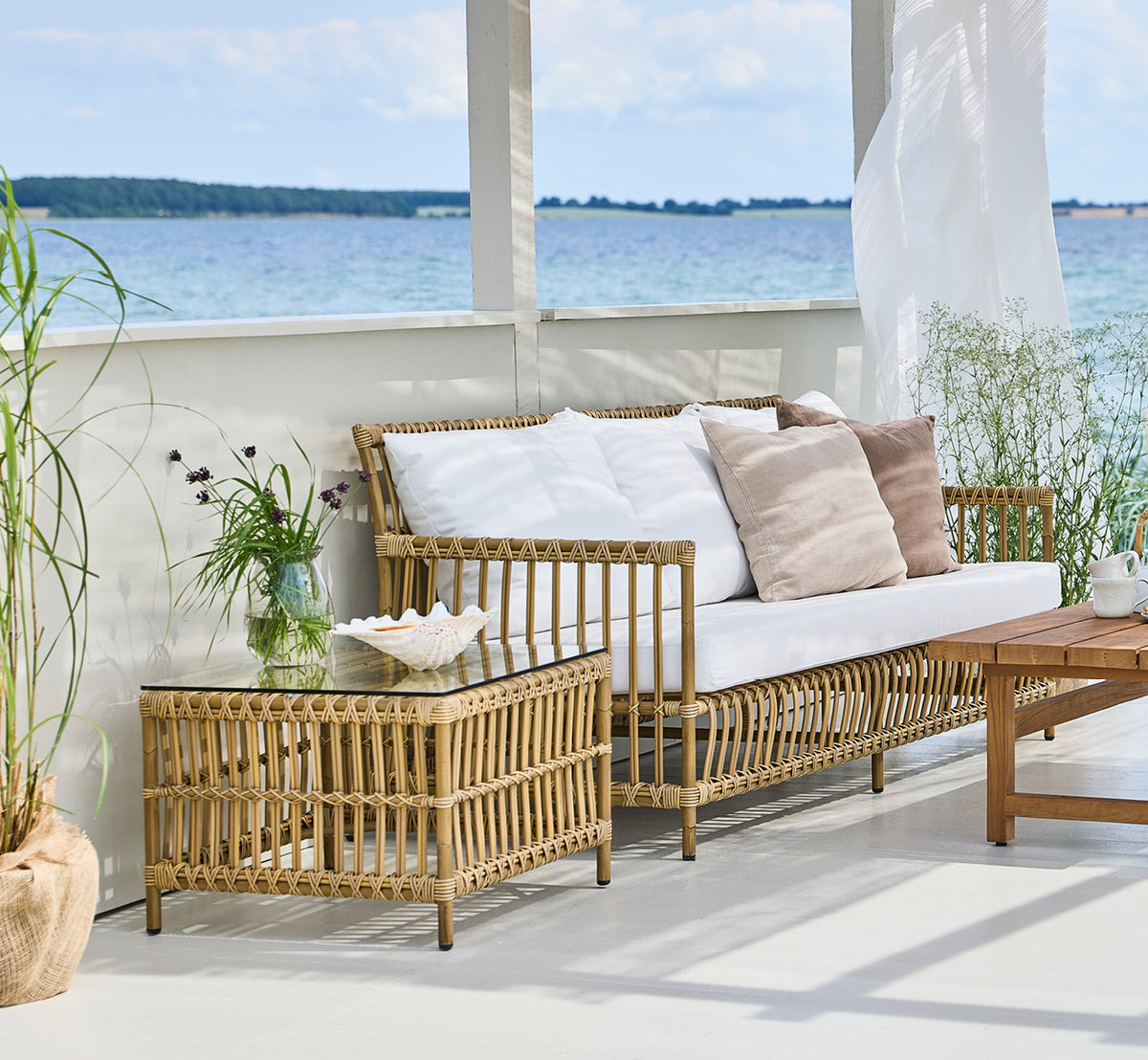 This is a lifestyle image of Caroline Exterior Table in antique Aluminium ArtFibre, on a waterside wooden deck with coastal greenery and sea views.