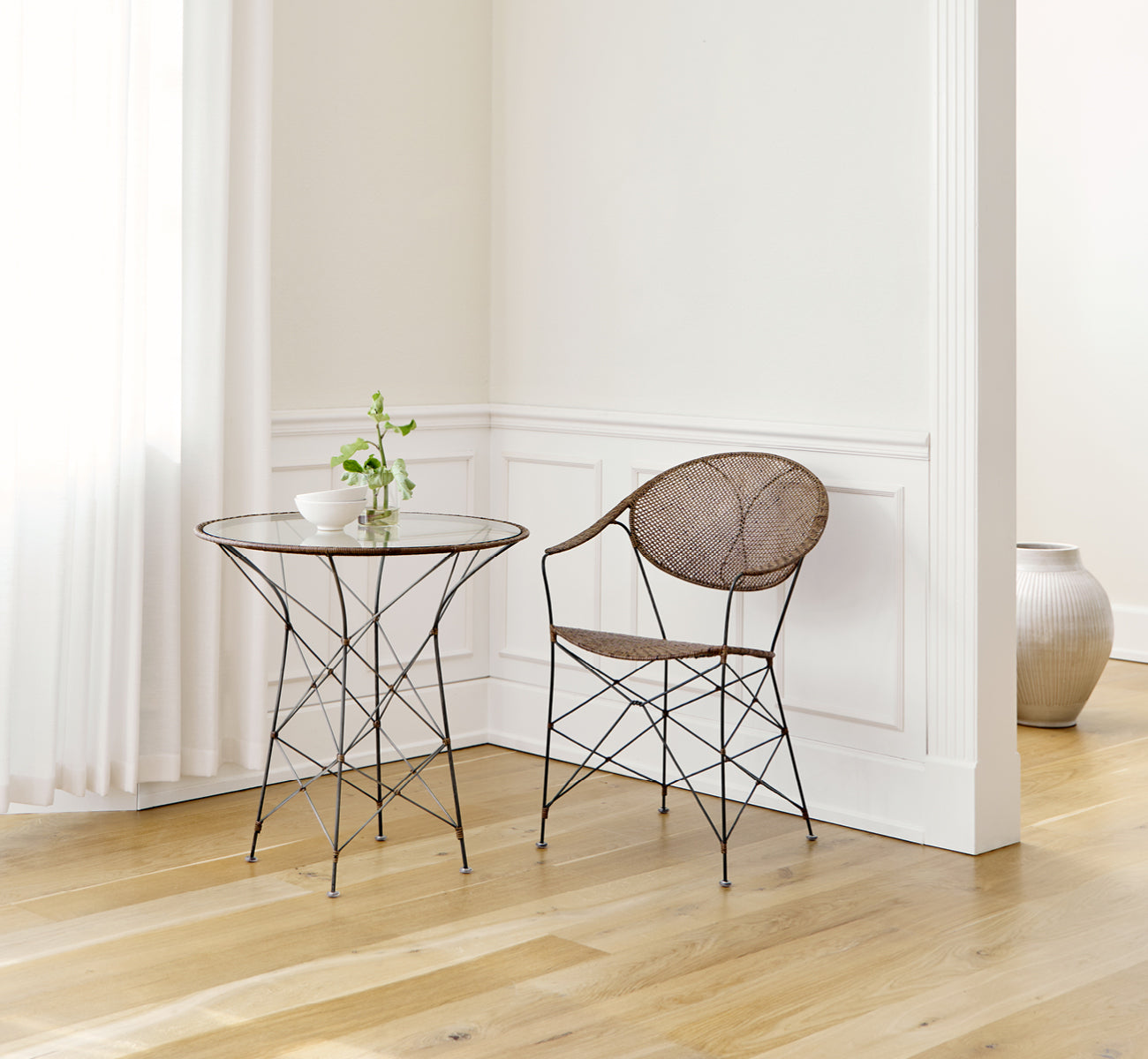 This is a lifestyle image of Funky Dining Chair in hazelnut rattan iron frame, in a bright interior with white panelled walls and herringbone parquet.