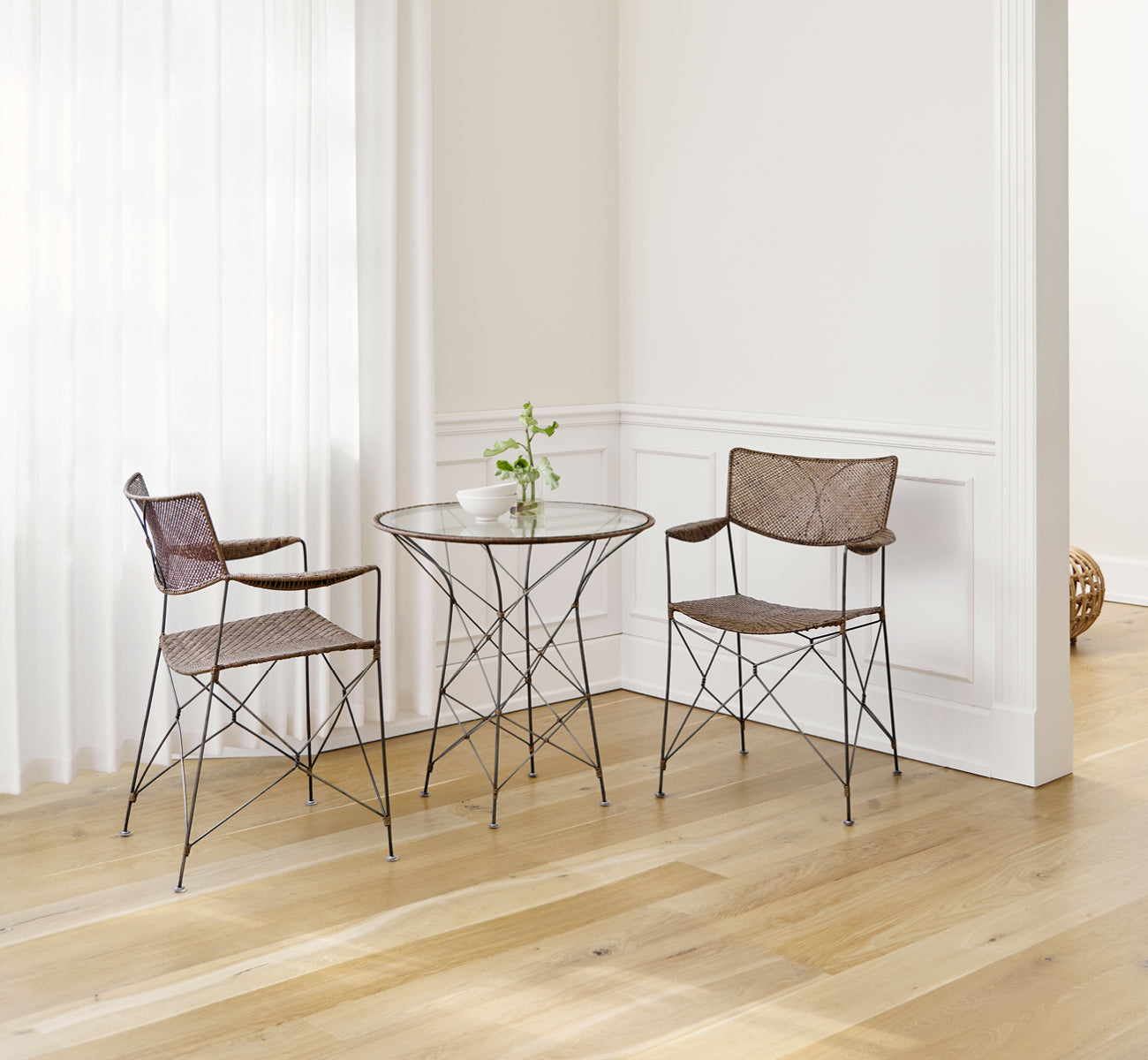 This is a lifestyle image of Rap Dining Chair in hazelnut rattan iron frame, in a bright interior with white panelled walls and herringbone parquet.