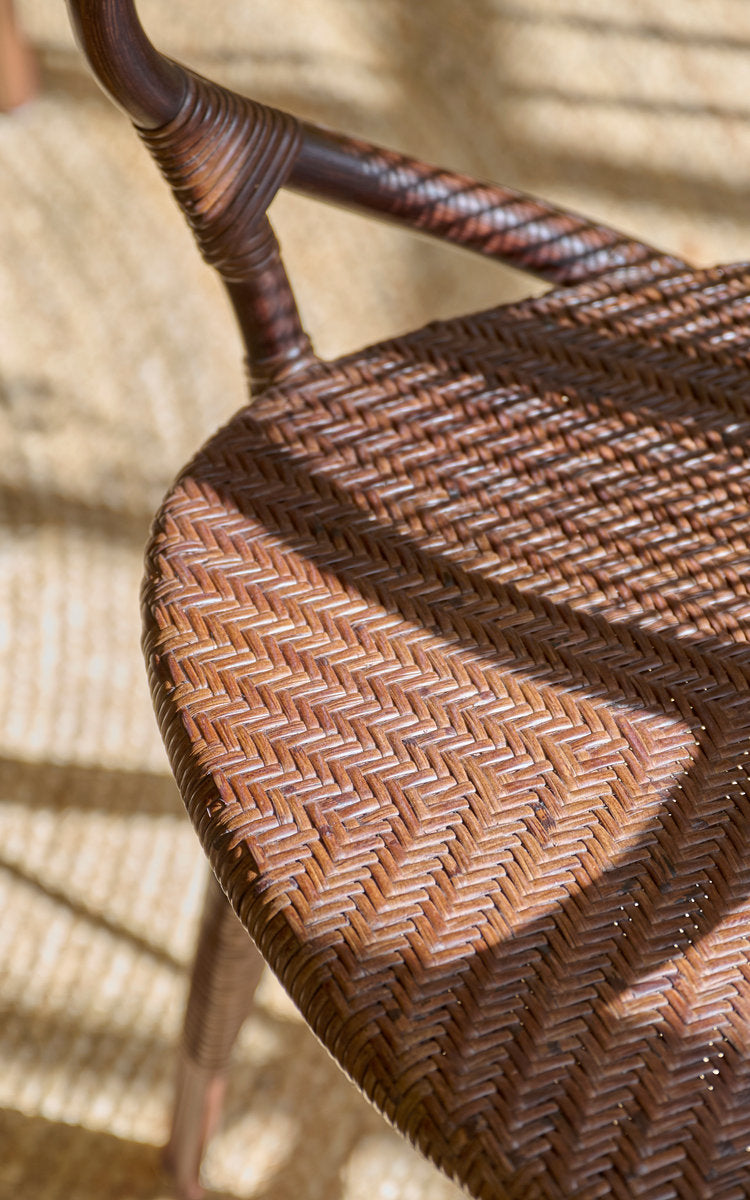 This is a close-up image of Rossini Dining Armchair in brown rattan. Herringbone seat weave and wrapped armrest in warm sunlight.