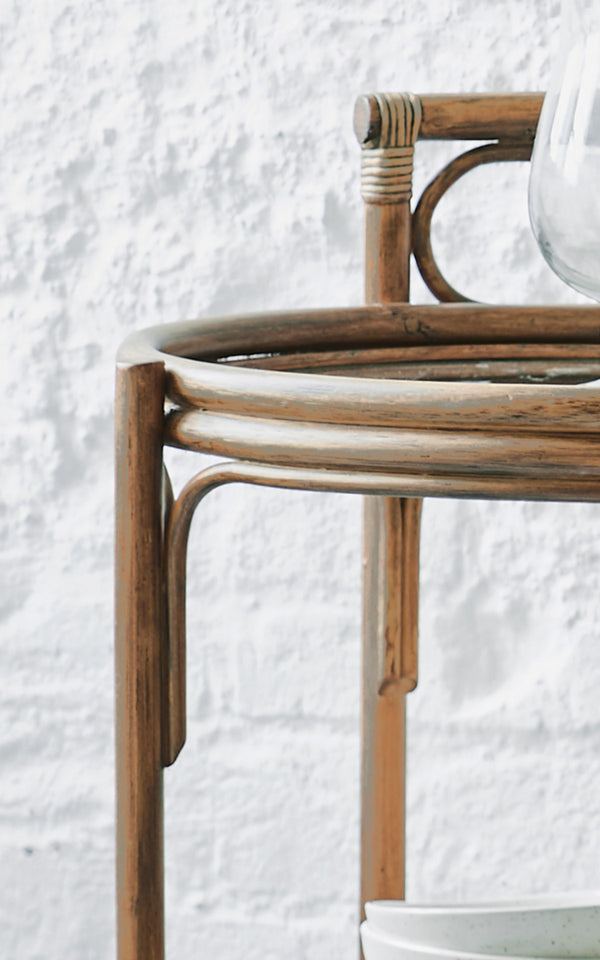 This is a close-up image of Romeo Trolley in antique rattan. Detail of the round rattan trolley frame against a white brick wall.