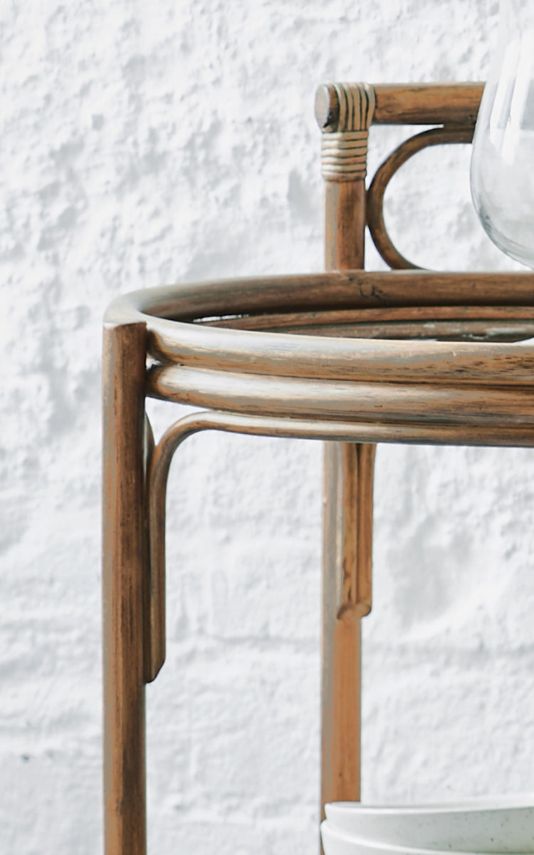 This is a close-up image of Romeo Trolley in antique rattan. Detail of the round rattan trolley frame against a white brick wall.