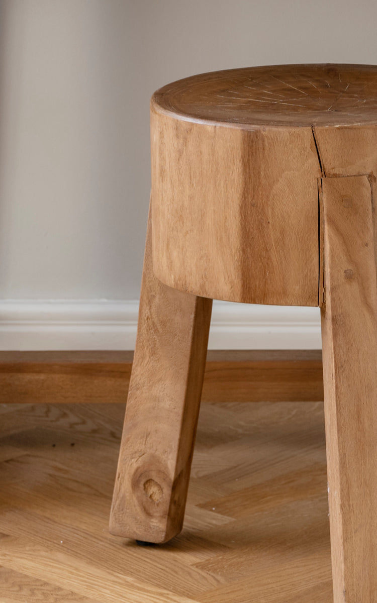 This is a close-up image of Roger Stool in teak indoor. Detail of the solid teak seat and three chunky angled legs.