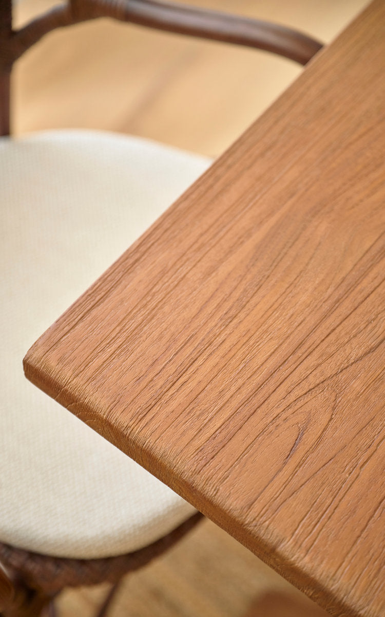 This is a close-up image of Hugo Teak Dining Table 140x90 cm in teak indoor. Smooth teak tabletop edge beside a rattan chair.