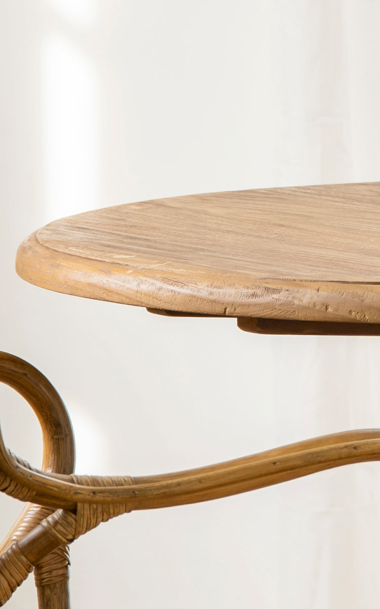 This is a close-up image of Michel Teak Dining Table Ø80 cm in teak indoor. The round tabletop edge and curved rattan pedestal base are shown.