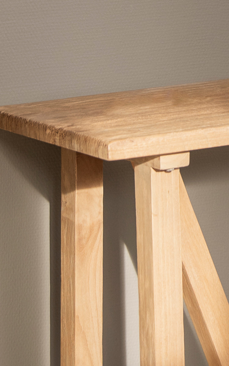 This is a close-up image of Lucas Teak Console in teak indoor. The solid teak surface and angled trestle legs show clean lines and wood tones.