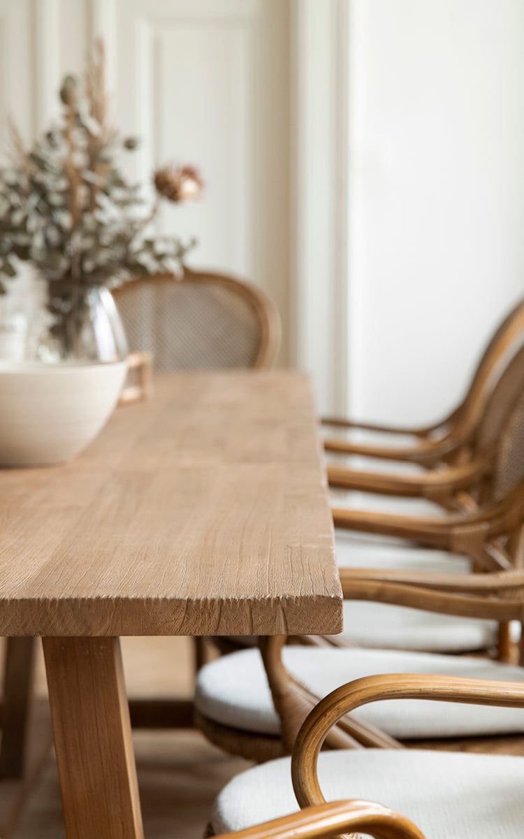 This is a close-up image of Lucas Teak Dining Table 300X100 cm in teak indoor. The solid teak edge is shown with rattan chairs in the background.
