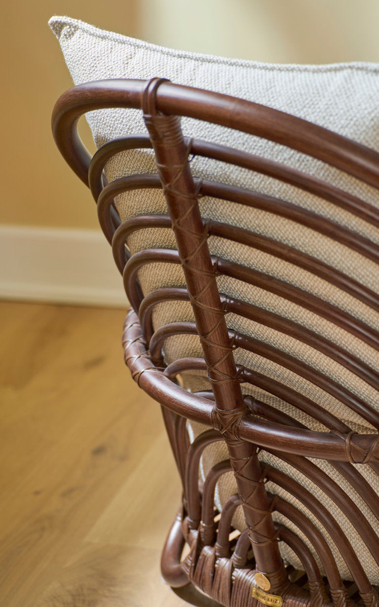 This is a close-up image of Charlottenborg Lounge Chair in brown rattan. Curved rattan back with linen cushion.