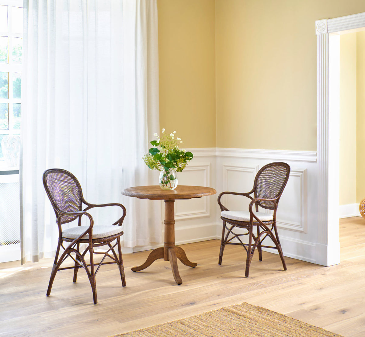 This is a lifestyle image of Rossini Dining Armchair in brown rattan. Pair by a teak café table and white paneled wall.