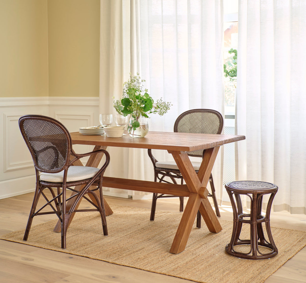This is a lifestyle image of Rossini Dining Armchair in brown rattan. Teak table, rattan stool and jute rug in a bright room.