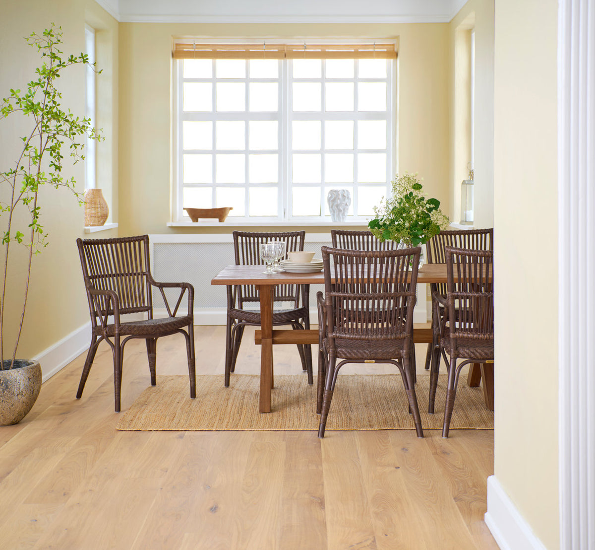 This is a lifestyle image of Piano Dining Chair in brown rattan. Around a teak dining table in a bright yellow room.