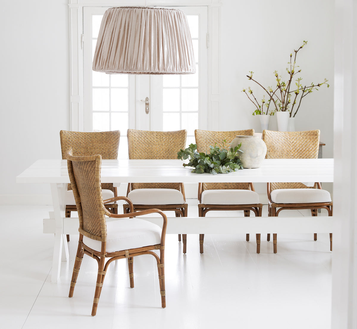 This is a lifestyle image of Melody Dining Armchair in antique rattan. Around a white table with linen pendant and spring branches.