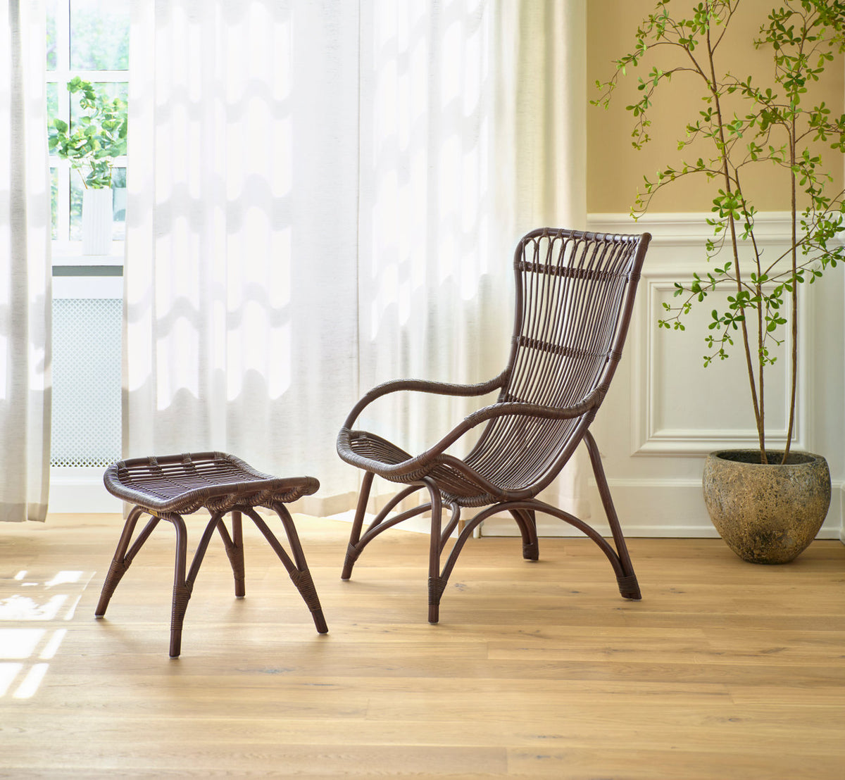 This is a lifestyle image of Monet Lounge Chair in brown rattan. With footstool and plant by sheer curtains.