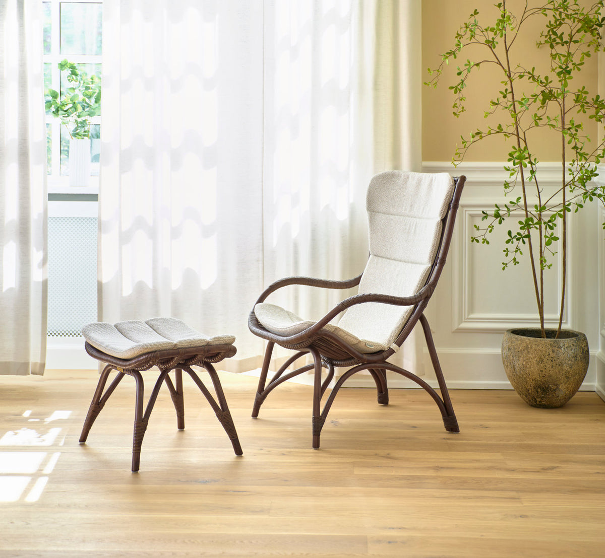 This is a lifestyle image of Monet Lounge Chair in brown rattan. With footstool, cushions and plant in a bright room.