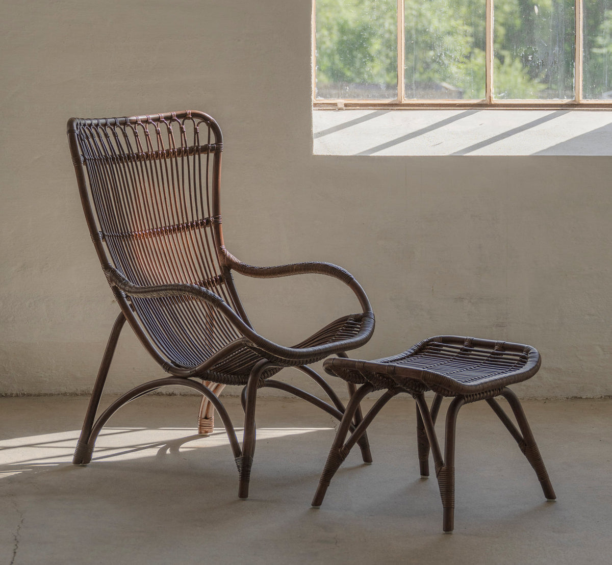 This is a lifestyle image of Monet Lounge Chair in brown rattan. With footstool in a sunlit room with concrete floor.