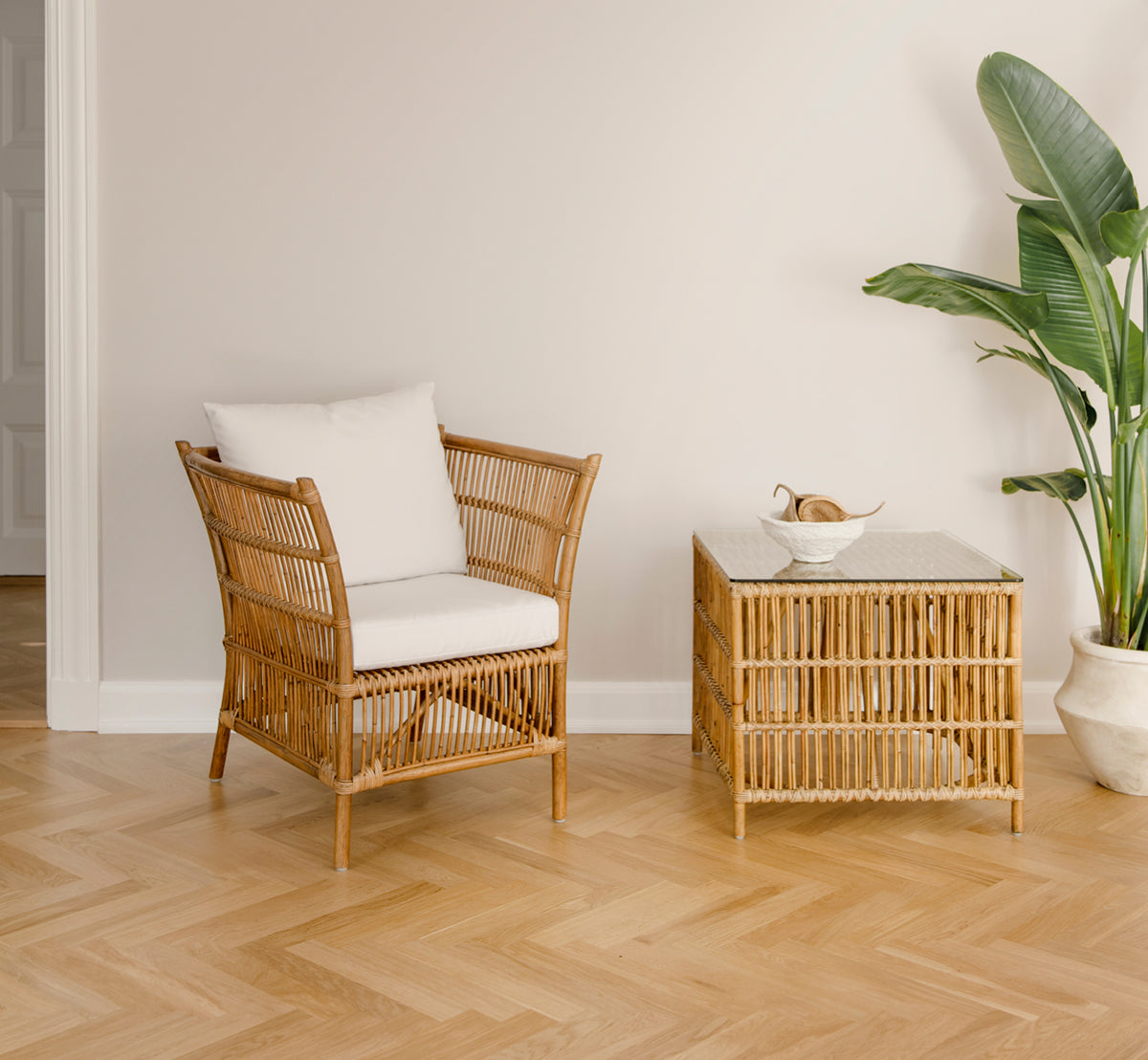 This is a lifestyle image of Donatello Lounge Chair in antique rattan, in a bright living room with herringbone parquet and lush greenery.