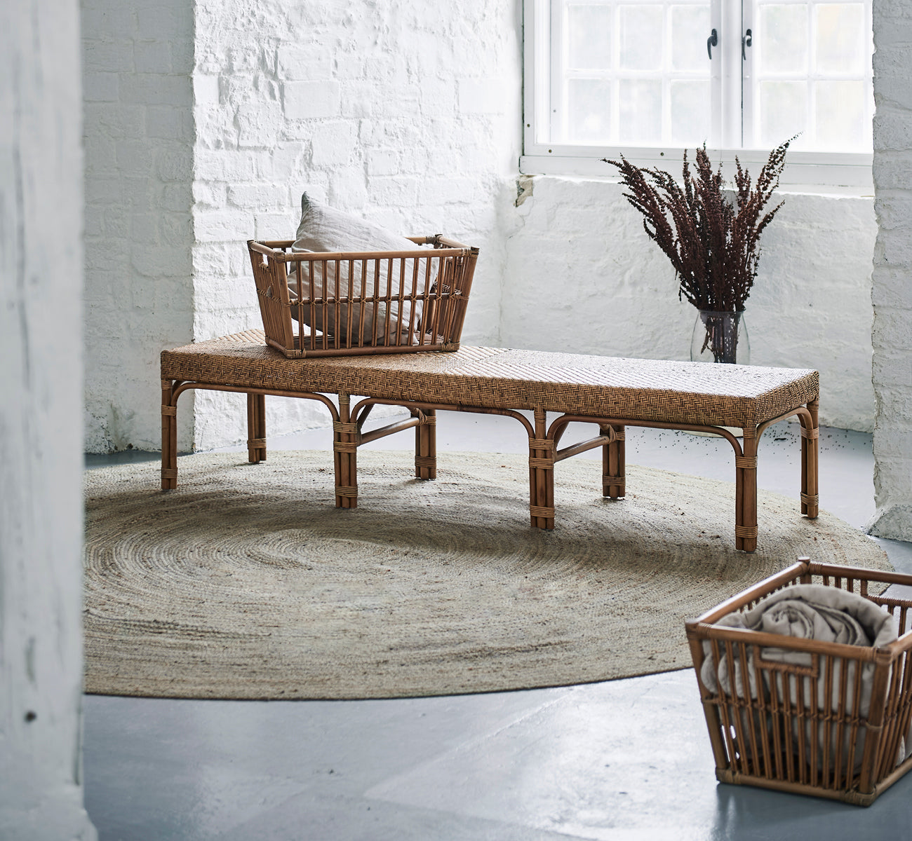 This is a lifestyle image of Marche Basket in antique rattan. Placed on a bench with cushions in a white brick interior with dried grasses.