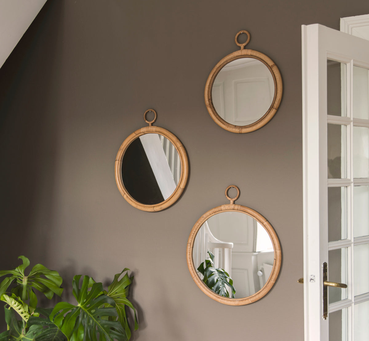 This is a lifestyle image of Ella Mirror O40cm in natural skin on rattan, on a taupe wall in a hallway with a monstera plant and white door.