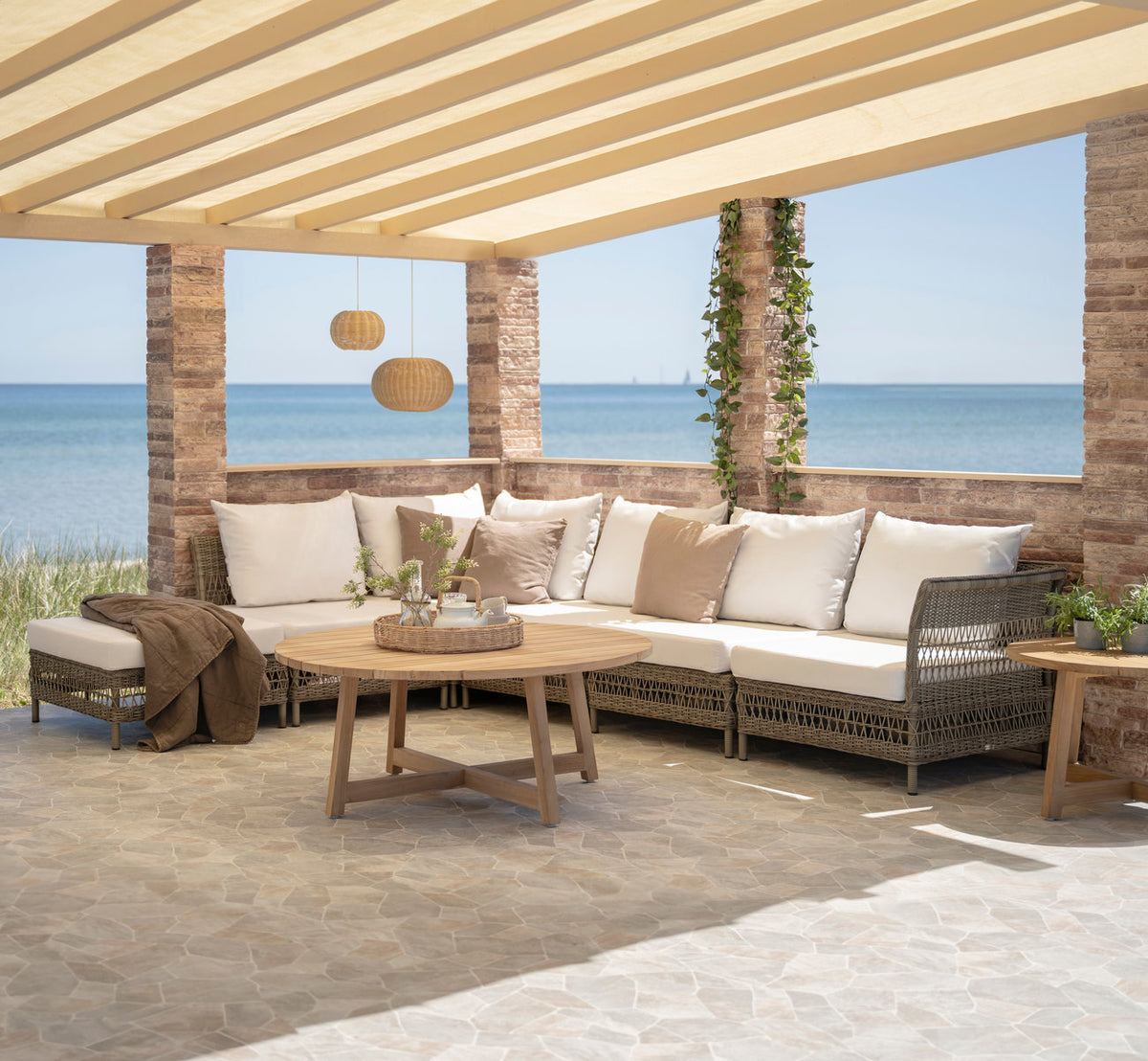 This is a lifestyle image of Mona Exterior Modular Sofa-Corner Module in antique grey artfibre aluminium. Under a pergola by the sea.