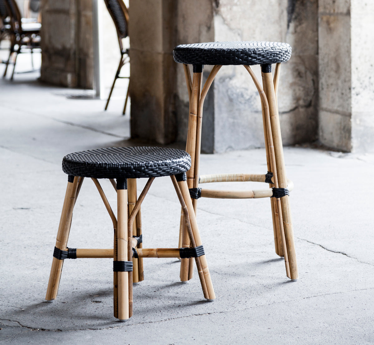 This is a lifestyle image of Simone Counter Stool in black ArtFibre Skin on Rattan frame. Placed with a stool by stone columns in a café setting.