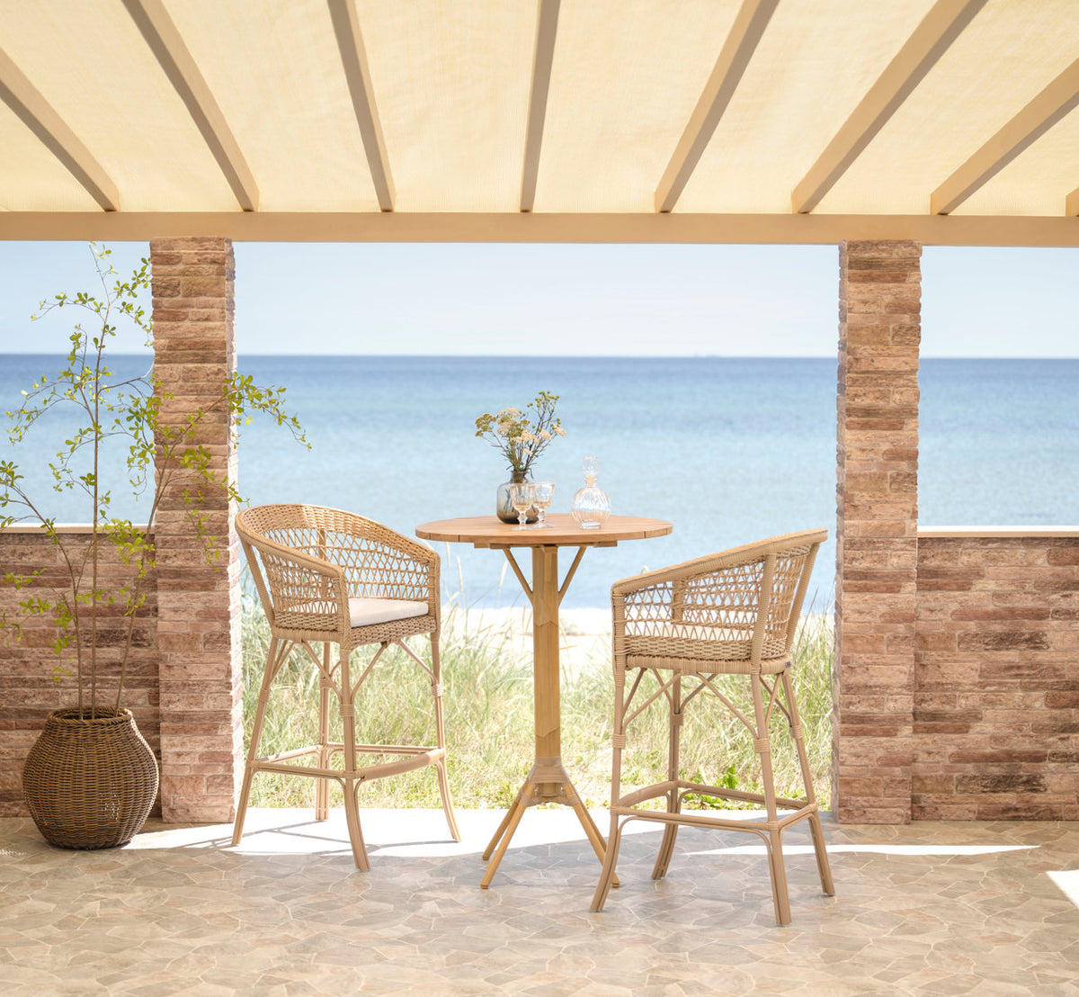 This is a lifestyle image of Emma Exterior Barstool in natural artfibre aluminium. Bar setting on a terrace by the sea.