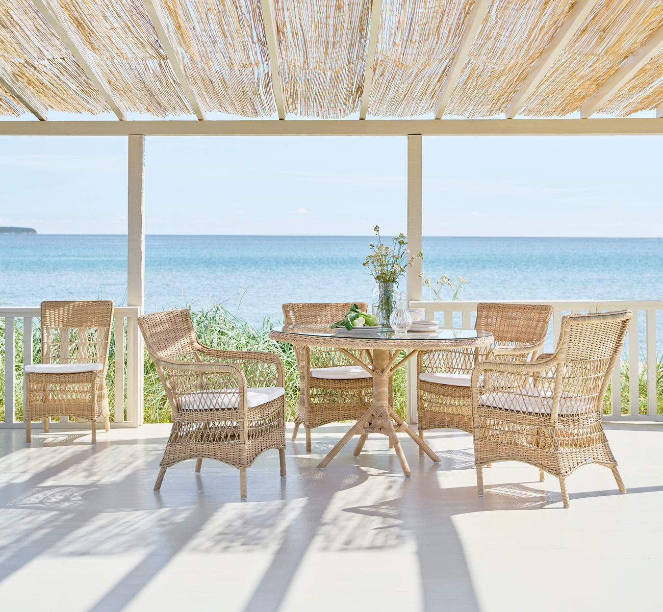This is a lifestyle image of Marie Exterior Side Chair in natural artfibre aluminium. Around a table on a shaded seaside veranda.