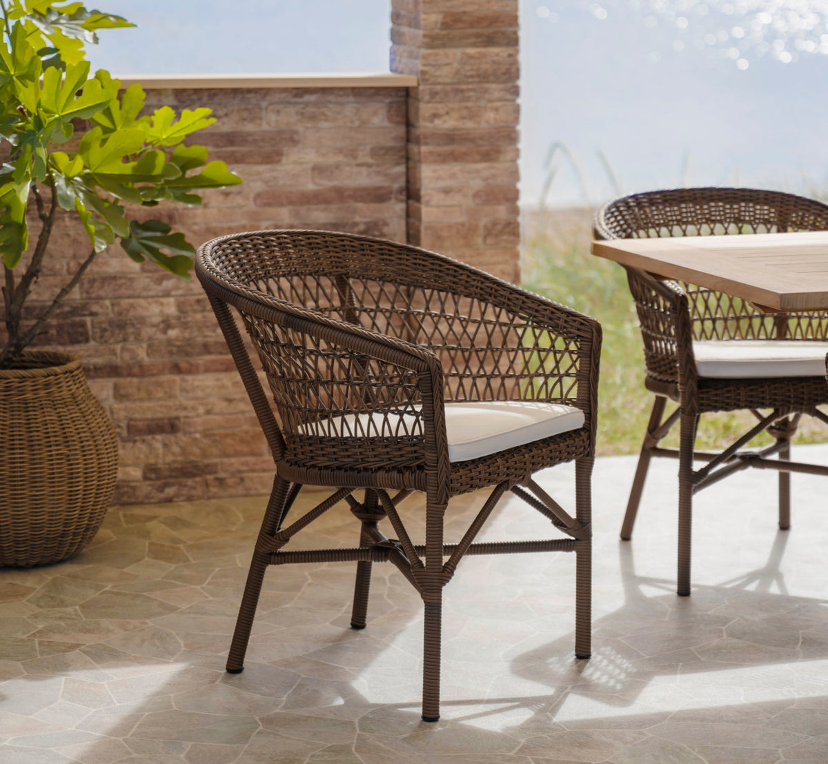 This is a lifestyle image of Emma Exterior Chair in brown artfibre aluminum. By a sunlit brick wall on a terrace.