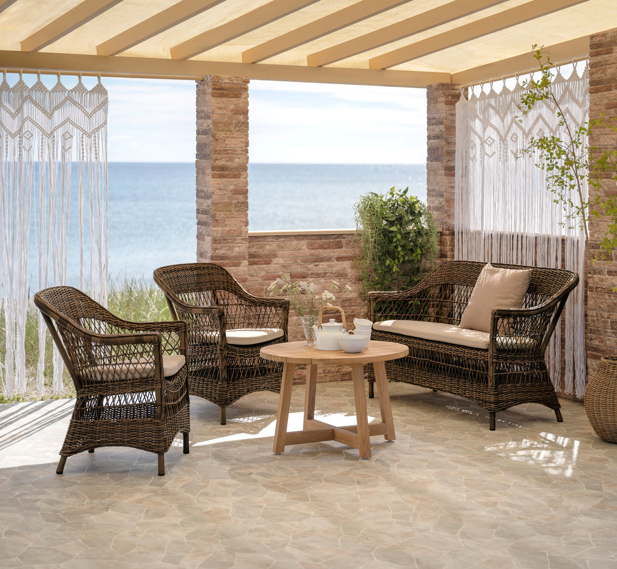 This is a lifestyle image of Charlot 2-Seater Exterior Sofa in brown artfibre aluminum. With teak coffee table and macramé curtains by the sea.