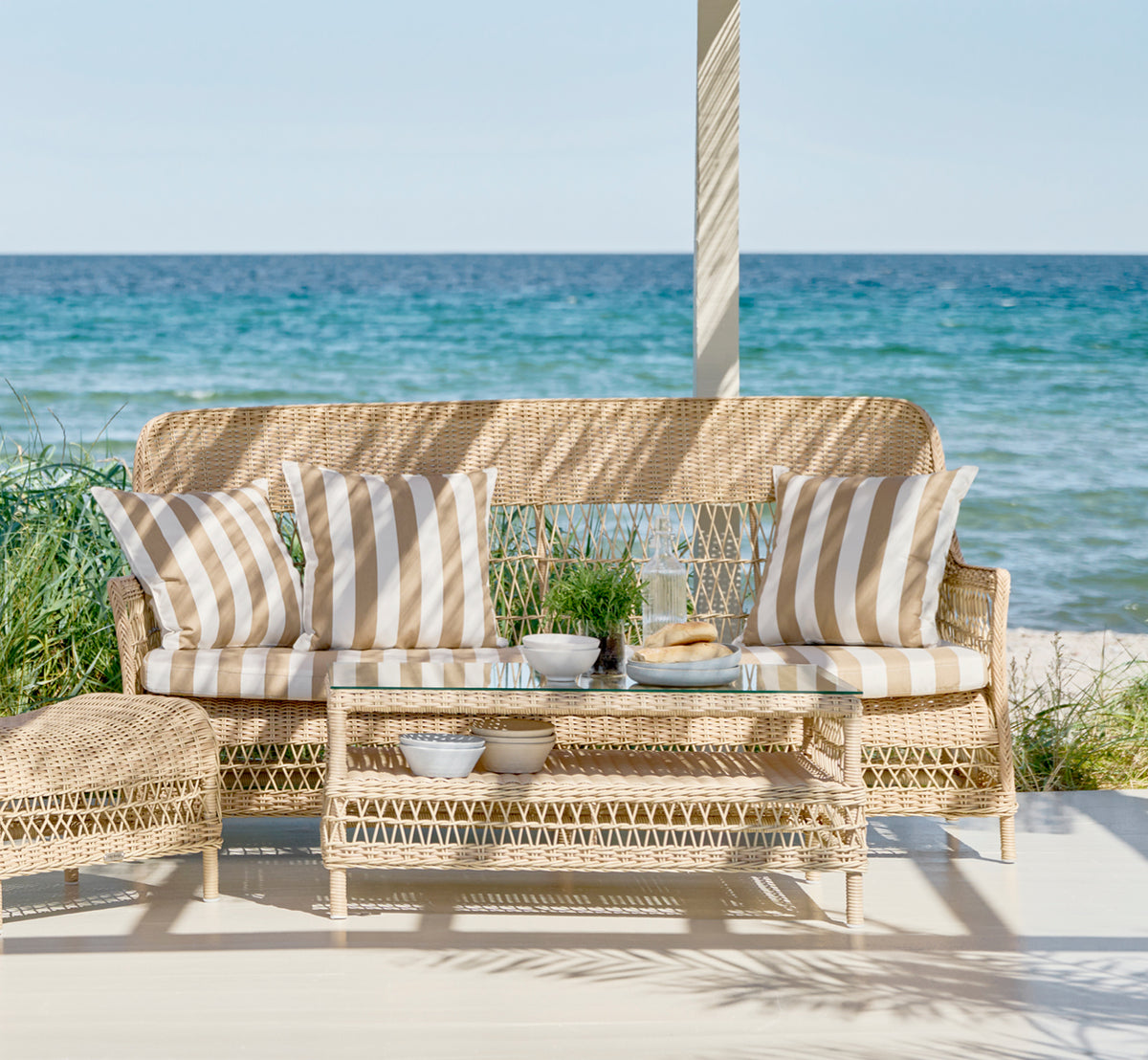 This is a lifestyle image of Dawn Sofa in natural ArtFibre Aluminium, on a coastal terrace with white wooden decking and sea views.