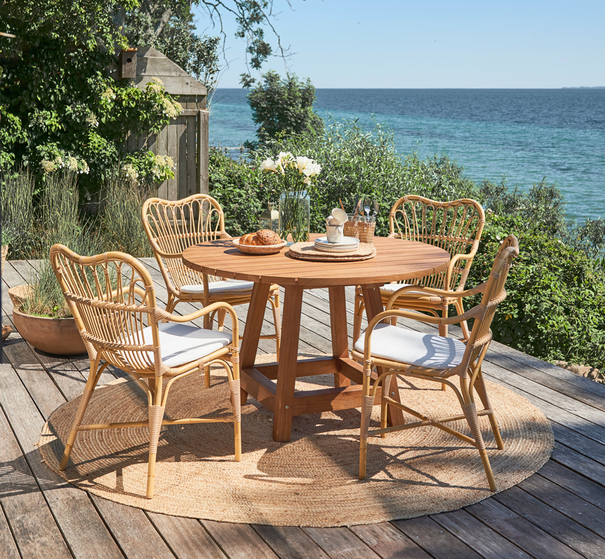 This is a lifestyle image of George Exterior Table Ø120 in outdoor Teak, on an outdoor garden patio with a white house facade and greenery.