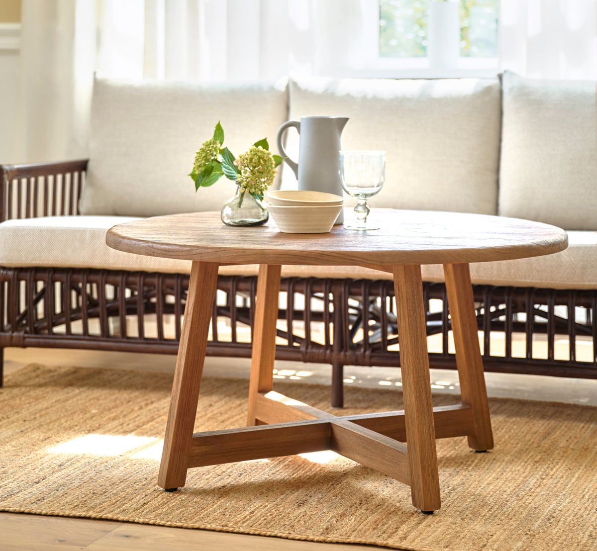 This is a lifestyle image of Aksel Coffee Table Ø100 cm in teak indoor. Before a rattan sofa on a jute rug.