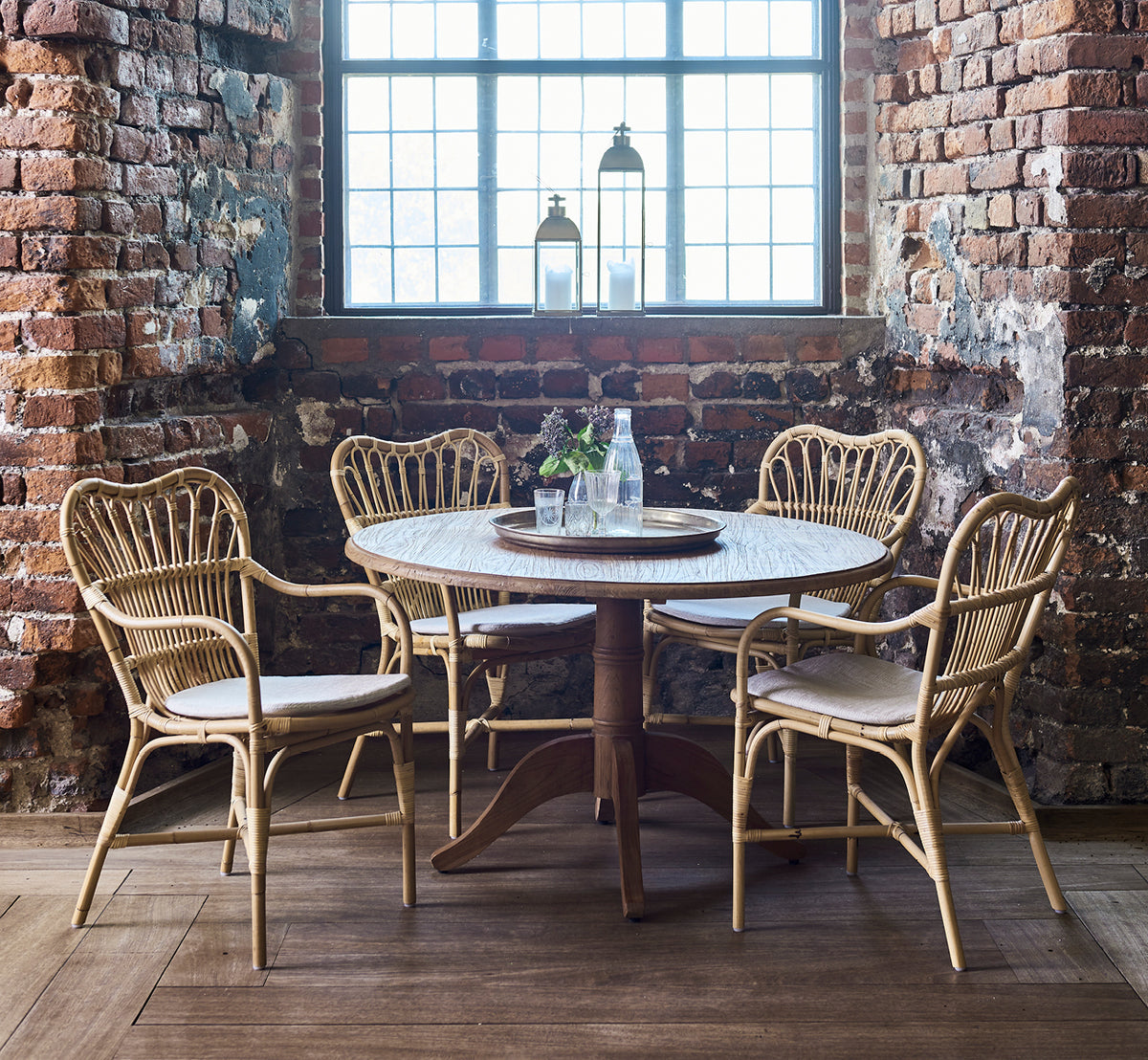 This is a lifestyle image of Michel Teak Dining Table Ø120 cm in teak indoor. Set in a raw brick loft with rattan chairs and lanterns on the sill.