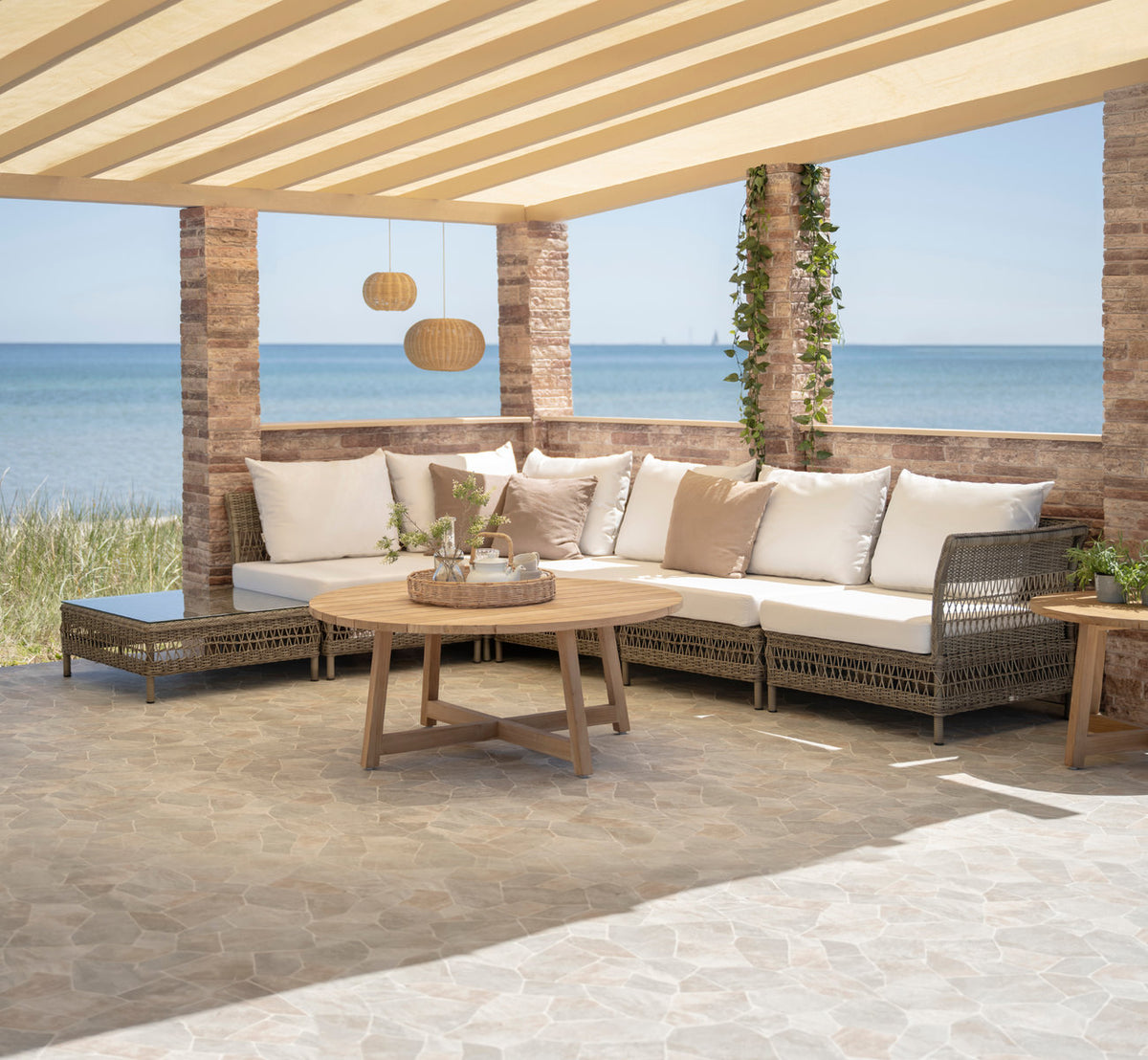 This is a lifestyle image of Victor Teak Lounge Table Ø120 cm in teak outdoor. Corner sofa with teak tables under a seaside pergola.