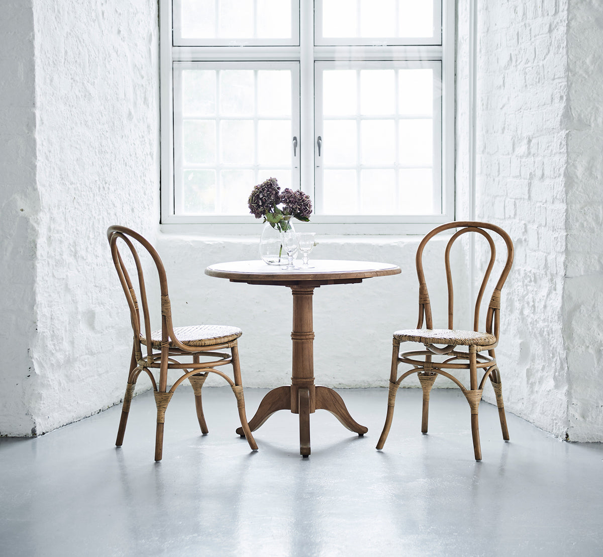 This is a lifestyle image of Michel Teak Dining Table Ø80 cm in teak indoor. Set with two chairs and flowers in a bright white brick interior.