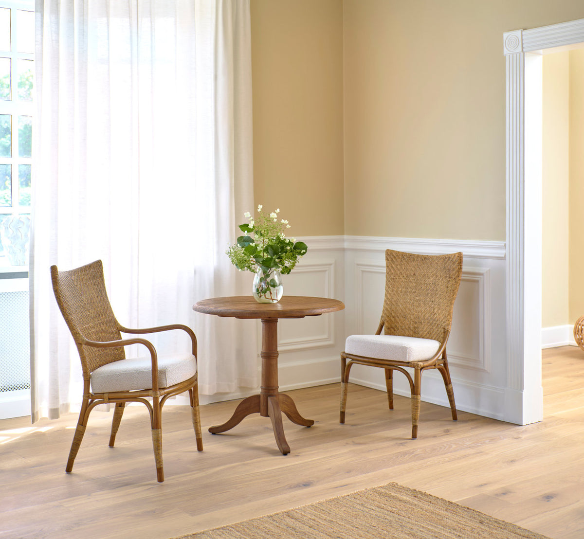 This is a lifestyle image of Michel Teak Dining Table Ø80 cm in teak indoor. With rattan chairs by white panels and curtains.
