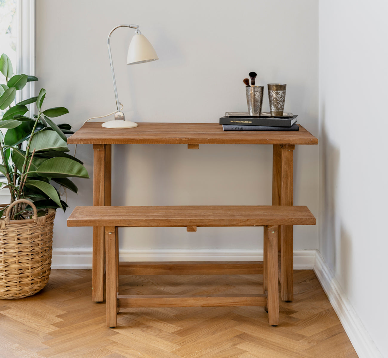 This is a lifestyle image of Lucas Teak Desk in teak indoor. Styled with a matching bench, desk lamp, and books in a cozy corner setting.