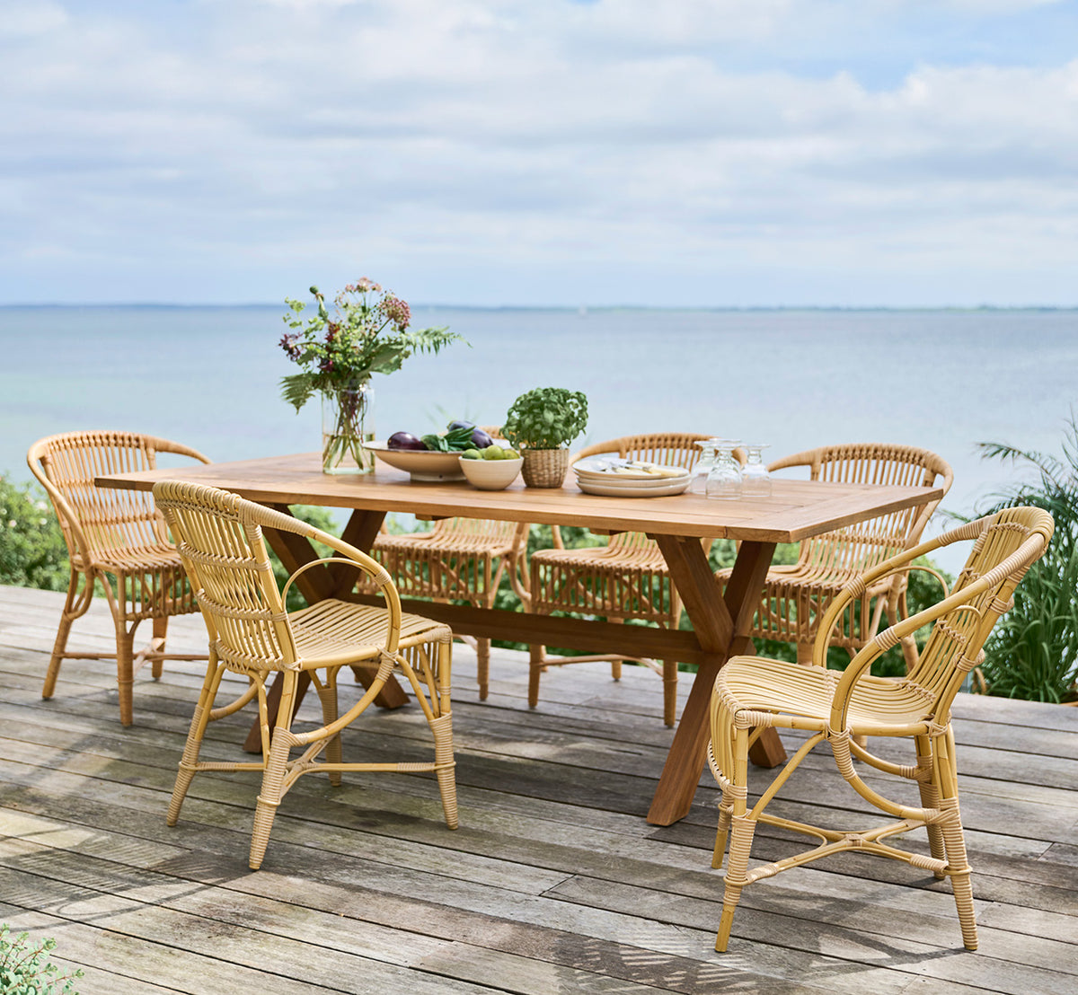 This is a lifestyle image of Colonial Table 200 in outdoor Teak, on a beachside terrace with white wooden decking and sea views.