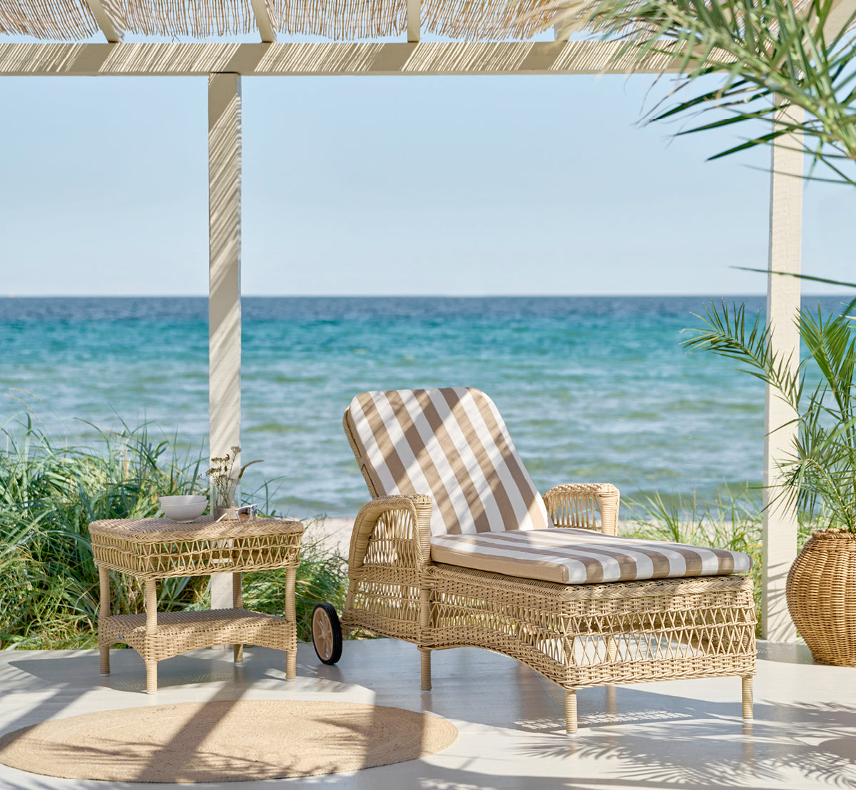 This is a lifestyle image of Susy Side Table in natural ArtFibre Aluminium, on a coastal terrace with sea views, beach grasses and white columns.