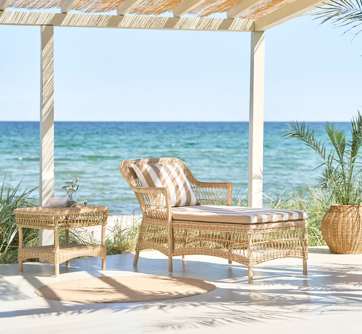 This is a lifestyle image of Olivia Exterior Sunbed in natural artfibre aluminium. In a lush garden with cushion and throw.