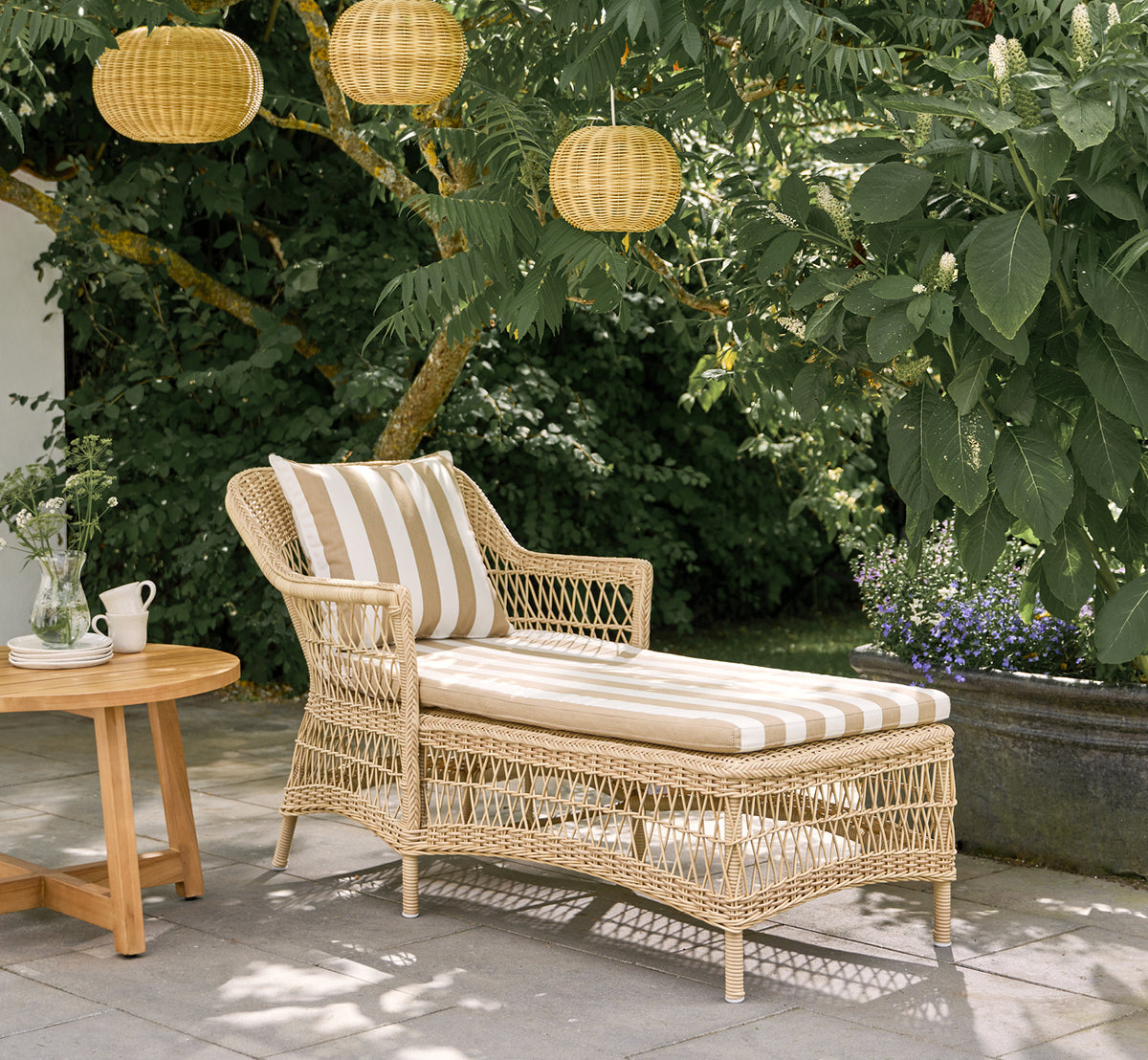 This is a lifestyle image of Olivia Exterior Sunbed in natural artfibre aluminium. On a patio with striped cushion and lanterns.