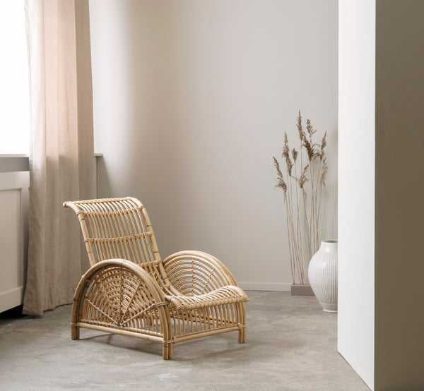 This is a lifestyle image of Paris Lounge Chair in polished natural rattan. Placed on a herringbone parquet floor beside a matching bench.