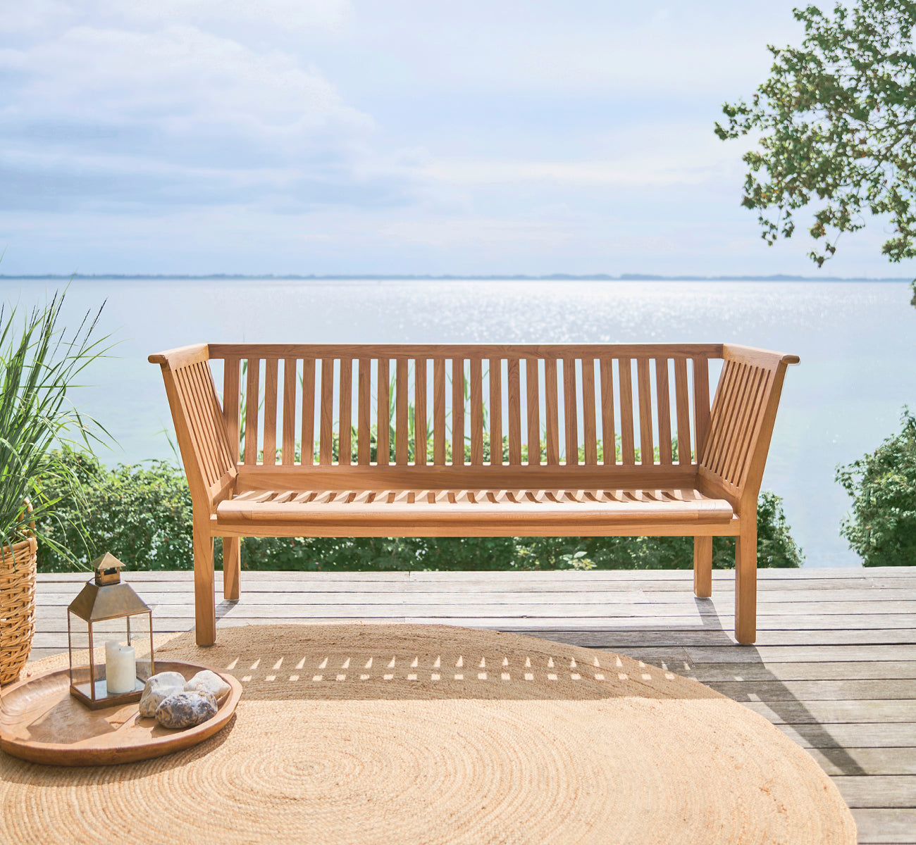 This is a lifestyle image of St Catherine Teak Bench in teak outdoor. Set on a wooden deck with sea view and a round jute rug.