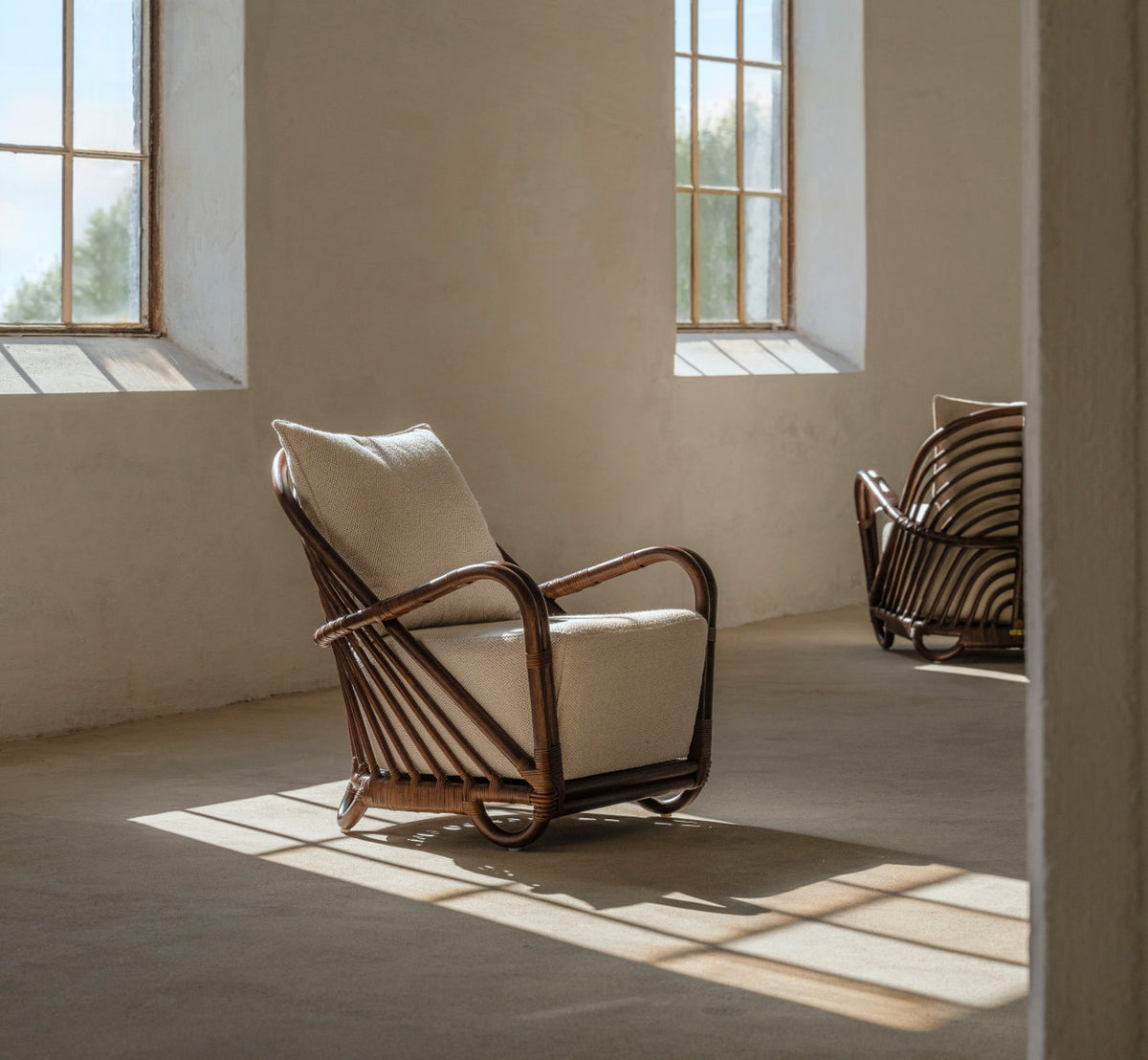 This is a lifestyle image of Charlottenborg Lounge Chair in brown rattan. With cushions in a sunlit room with concrete floor.