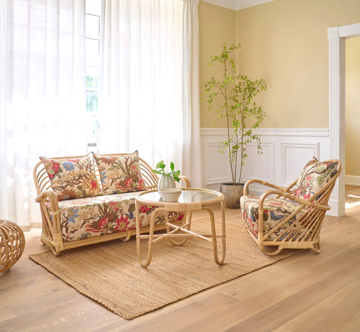 This is a lifestyle image of Charlottenborg Lounge Chair in polished natural rattan. With floral cushions and coffee table on a jute rug.