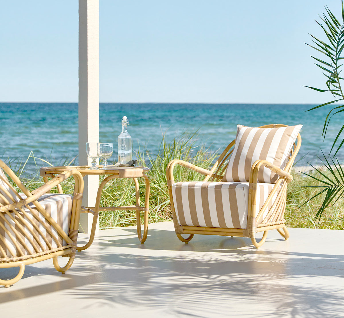 This is a lifestyle image of Charlottenborg Exterior Lounge Chair in natural Aluminium ArtFibre, on an outdoor wooden deck with flowers and sea views.