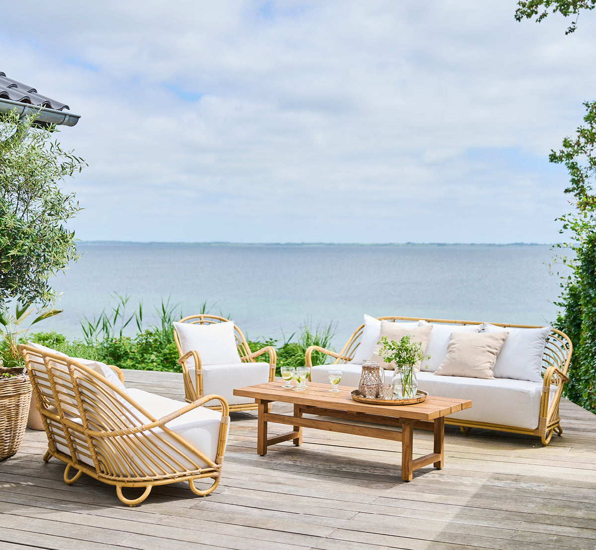 This is a lifestyle image of Charlottenborg Exterior Lounge Chair in natural Aluminium ArtFibre, on an outdoor wooden deck with flowers and sea views.