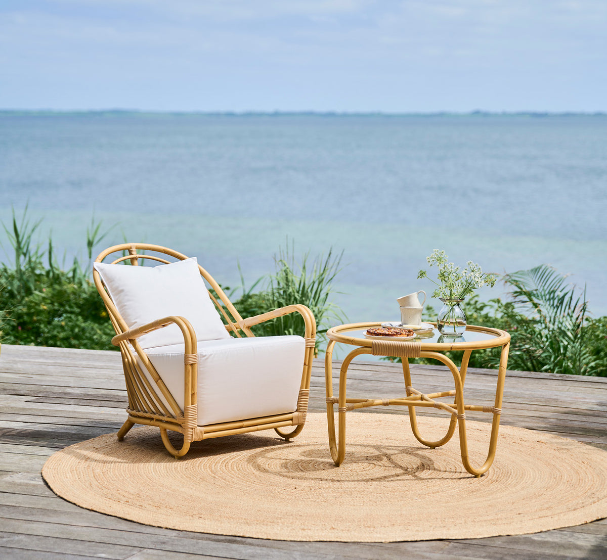 This is a lifestyle image of Charlottenborg Exterior Coffee Table in natural Aluminium ArtFibre, on an outdoor wooden deck with coastal greenery and.